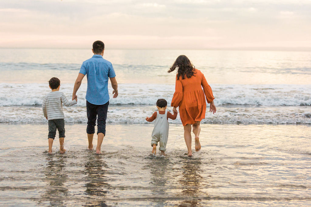 Founder family photo (mom, dad, two boys) with backs to camera stepping into the ocean